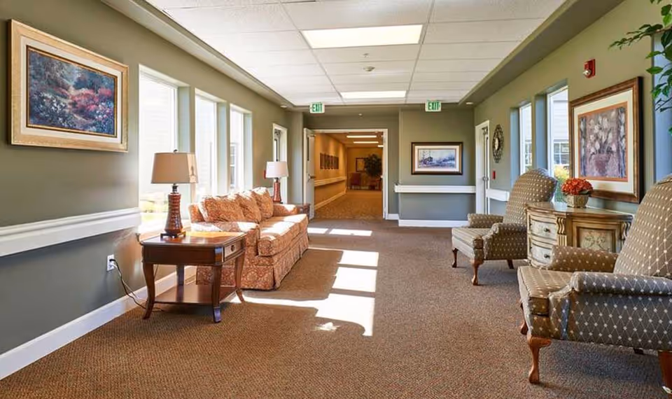 A well-lit hallway in an assisted living facility with large windows on one side letting in natural light. The hallway features a patterned sofa with two table lamps on a side table, two upholstered armchairs, framed artwork on the walls, and a small decorative table with a flower arrangement. The carpet is brown, and the walls are painted a muted green color. There are exit signs above the doors at the end of the hallway.