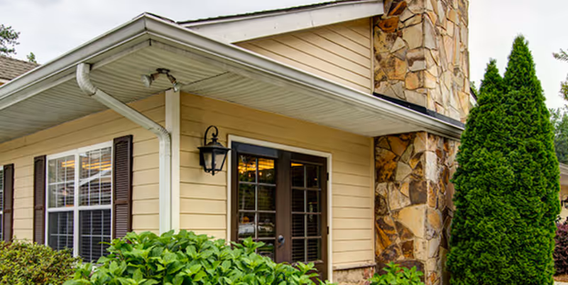Exterior corner view of a building with beige siding, a stone chimney, dark brown framed windows and door, surrounded by green bushes and a tall conifer tree.