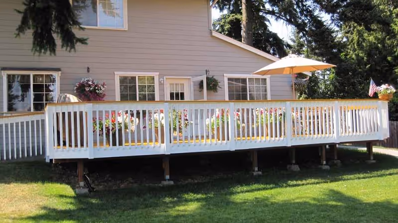 A raised wooden deck attached to a beige house with white trim. The deck has white railings and is decorated with various potted flowers and plants. There is a large beige patio umbrella providing shade over part of the deck. The house has several windows and a door leading to the deck. The surrounding area includes green grass and tall trees in the background.