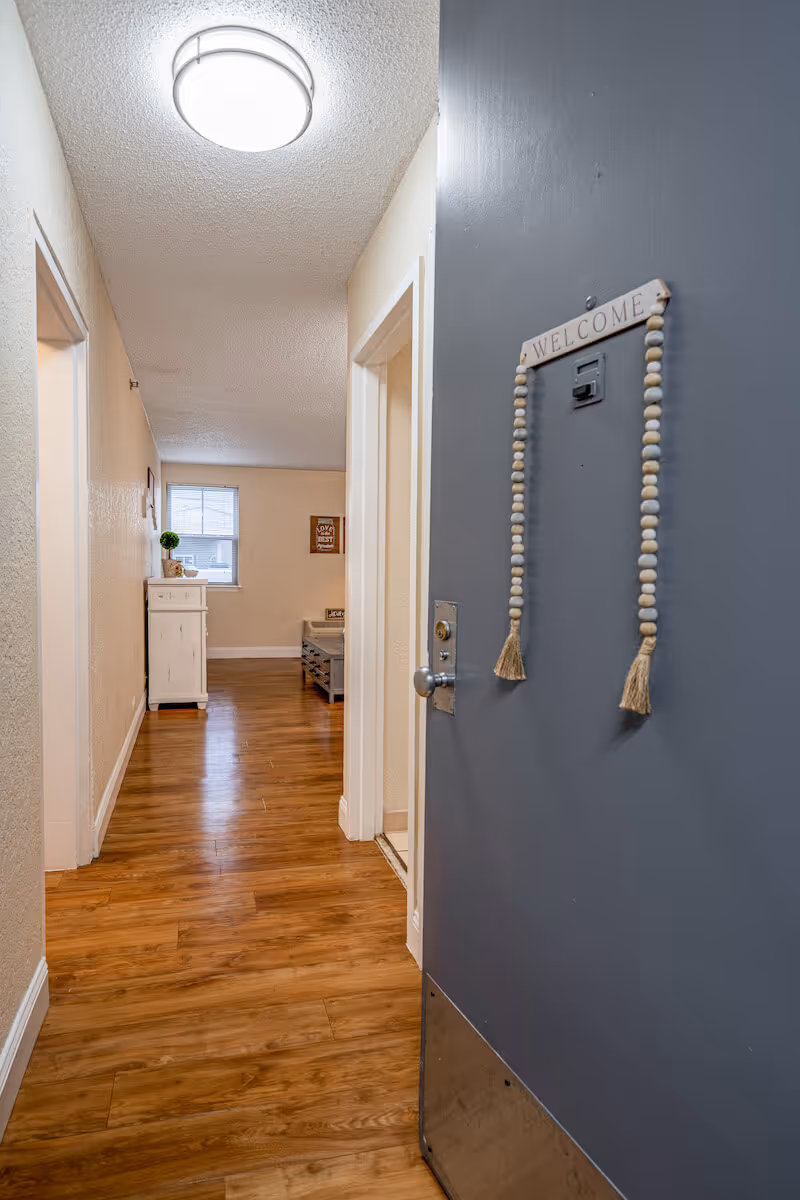 View looking down a hallway with wooden flooring inside a senior living facility. The open gray door on the right has a decorative 'WELCOME' sign with hanging beads. The hallway leads to a room with a window, a small white cabinet, and a bench. The walls are light-colored and there is a round ceiling light fixture.