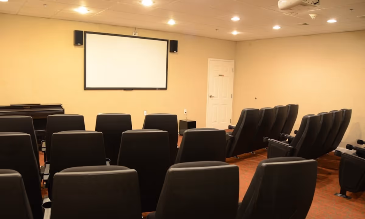 A small theater or media room with multiple rows of black cushioned chairs facing a large blank projection screen mounted on a beige wall. There is a piano on the left side of the room and a closed white door on the right wall. The ceiling has recessed lighting.