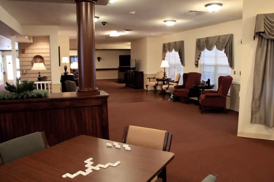 Interior view of a senior living community common area with a wooden table in the foreground featuring a game of dominoes, upholstered chairs, a wooden column, lamps, windows with drapes, and a carpeted floor.