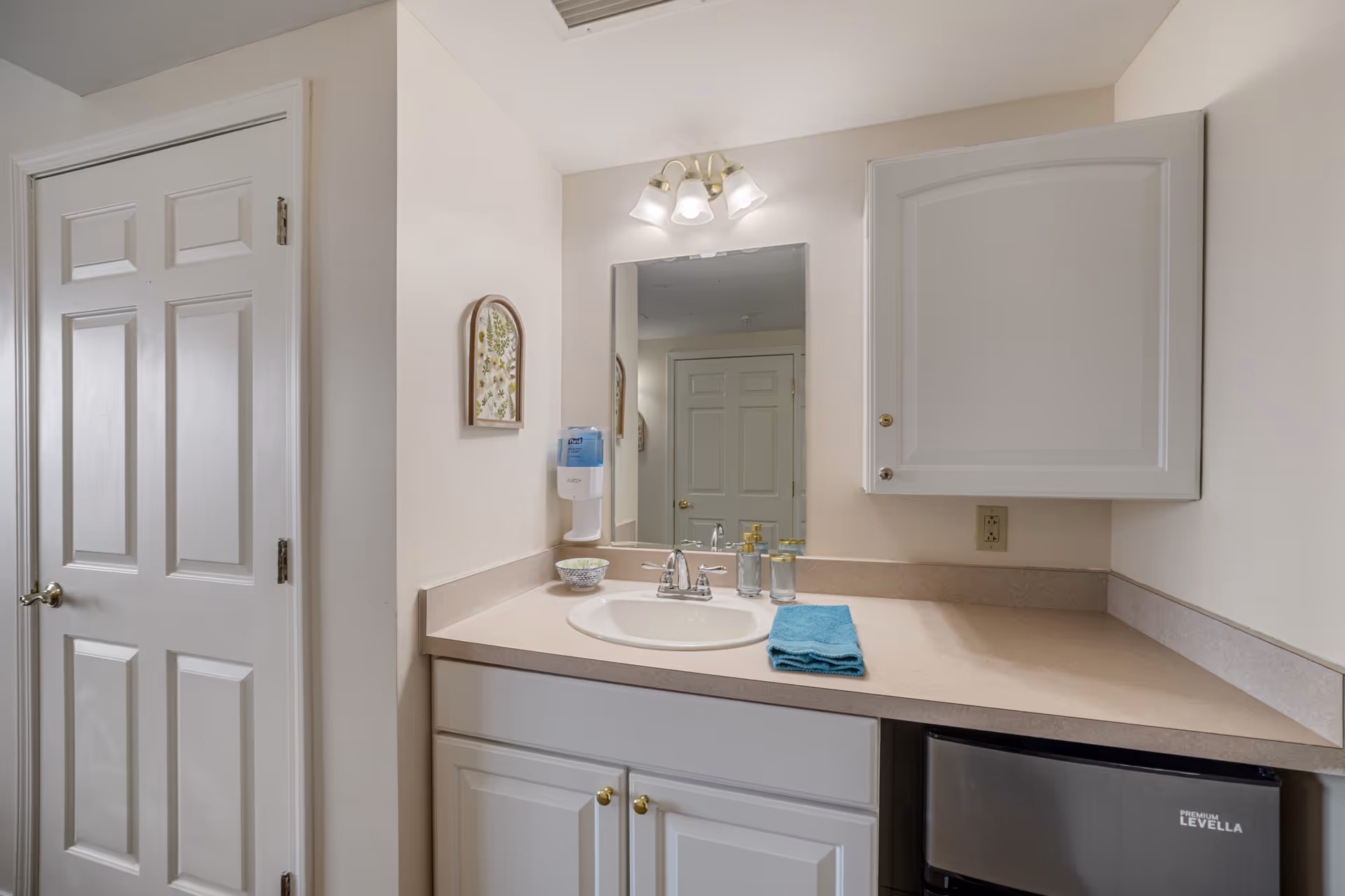 A bathroom vanity area with a beige countertop, white sink, and white cabinets below. Above the sink is a rectangular mirror with a three-light fixture. A blue hand towel, soap dispenser, and small bowl are on the countertop. To the right, there is a small black refrigerator labeled 'Premium Levella'. A white cabinet is mounted on the wall above the refrigerator. A closed white door is visible to the left, and a small framed botanical artwork hangs on the wall next to the mirror.
