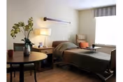 A well-lit bedroom in a healthcare facility featuring a single bed with green bedding, a bedside table with a lamp and picture frame, a comfortable chair with an orange cushion near a large window with blinds, and a round table with a vase of flowers and chairs.