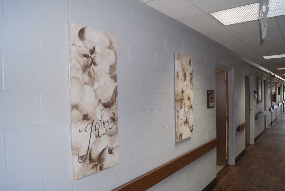 Long interior hallway with floral wall art, a wooden handrail, doors and fluorescent ceiling lights.