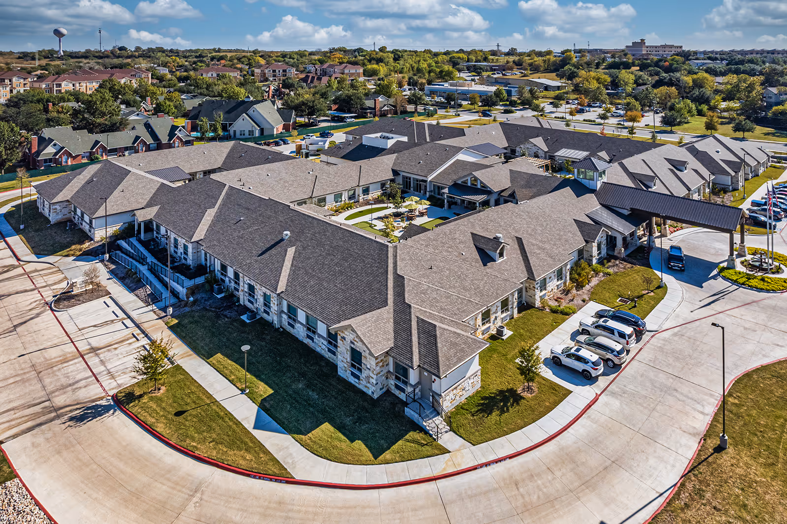 Aerial view of a large senior living facility with multiple connected buildings featuring stone and brick exteriors and gray shingled roofs. The facility has a covered entrance with a driveway and several parked cars. Surrounding the buildings are landscaped lawns, trees, and a courtyard with outdoor seating and umbrellas. Residential neighborhoods and other buildings are visible in the background under a partly cloudy sky.