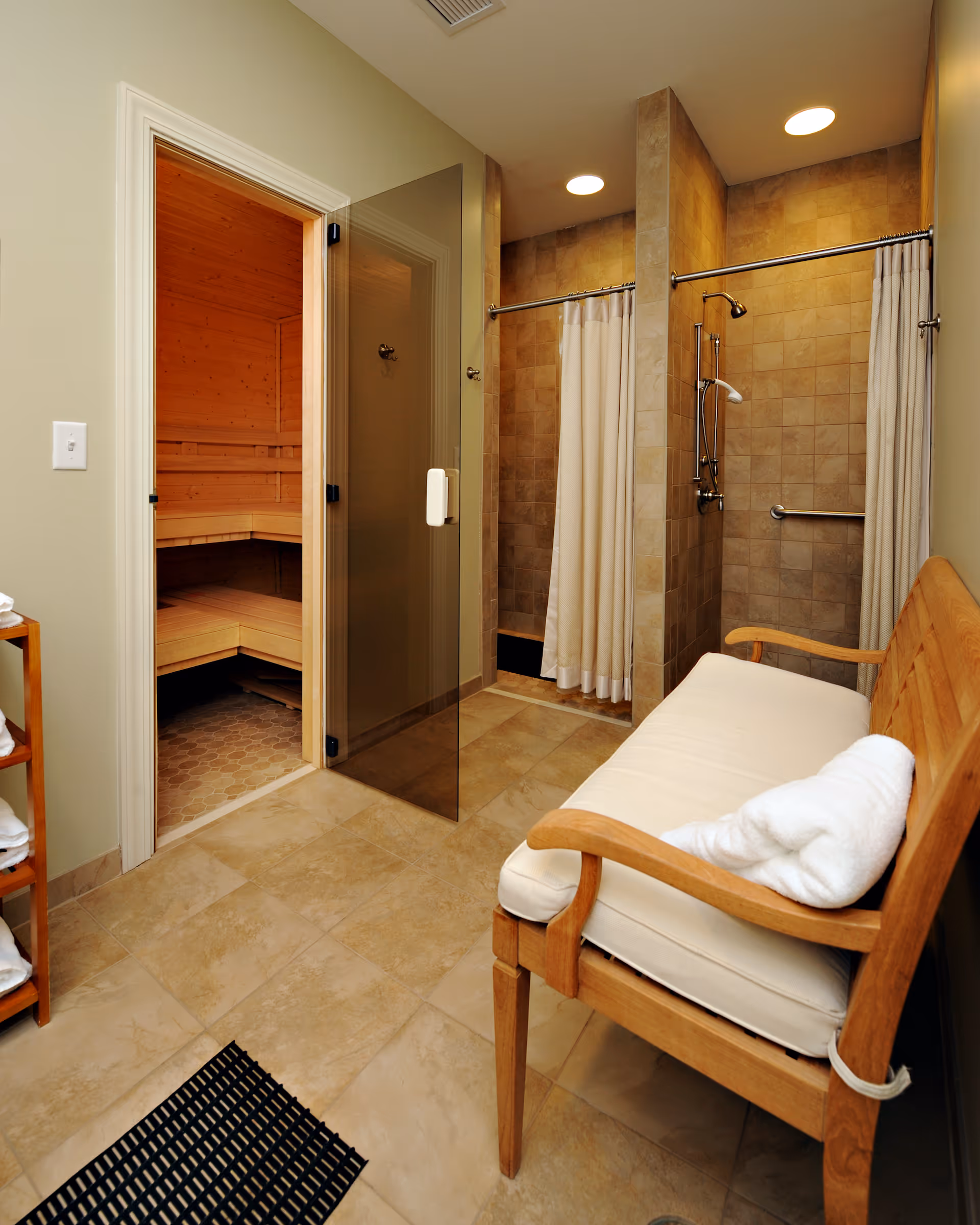 A clean and well-lit spa room featuring a wooden bench with a white cushion and a rolled towel on it. To the left, there is a glass door leading to a wooden sauna. On the right side, there are two tiled shower stalls with white curtains and chrome shower fixtures. The floor is tiled in a beige tone, and there is a small wooden shelf with folded white towels.