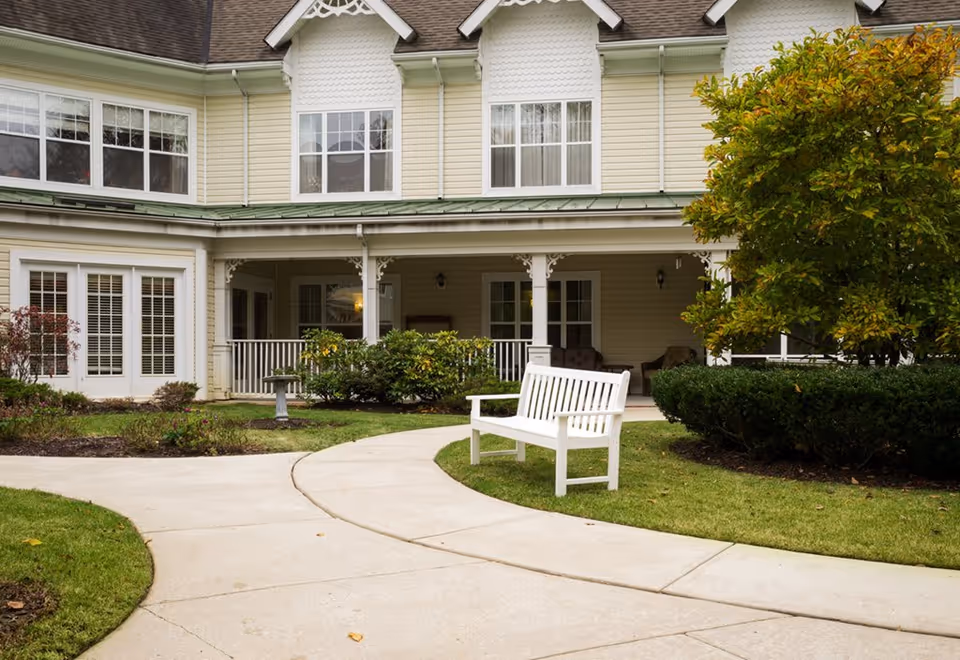 Outdoor view of a senior living facility with a curved concrete pathway, a white bench on the grass, bushes, and a tree in front of a two-story building with beige siding and white-framed windows.