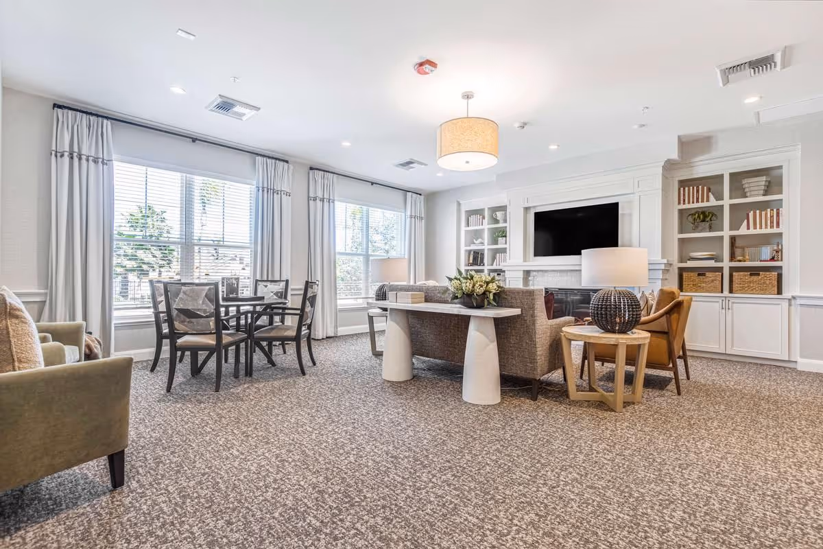 Bright communal living room with sofas, chairs, a dining table, built-in shelving and a large flat-screen TV in front of large windows.