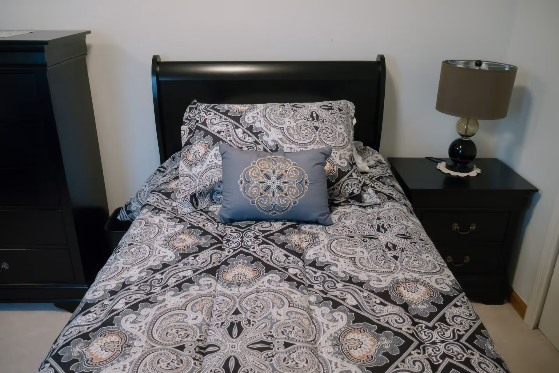A neatly made single bed with a black wooden headboard, patterned bedding in shades of gray, black, and beige, and a decorative blue pillow with an embroidered design. To the right of the bed is a black nightstand with two drawers and a lamp with a dark shade. To the left is a tall black dresser.