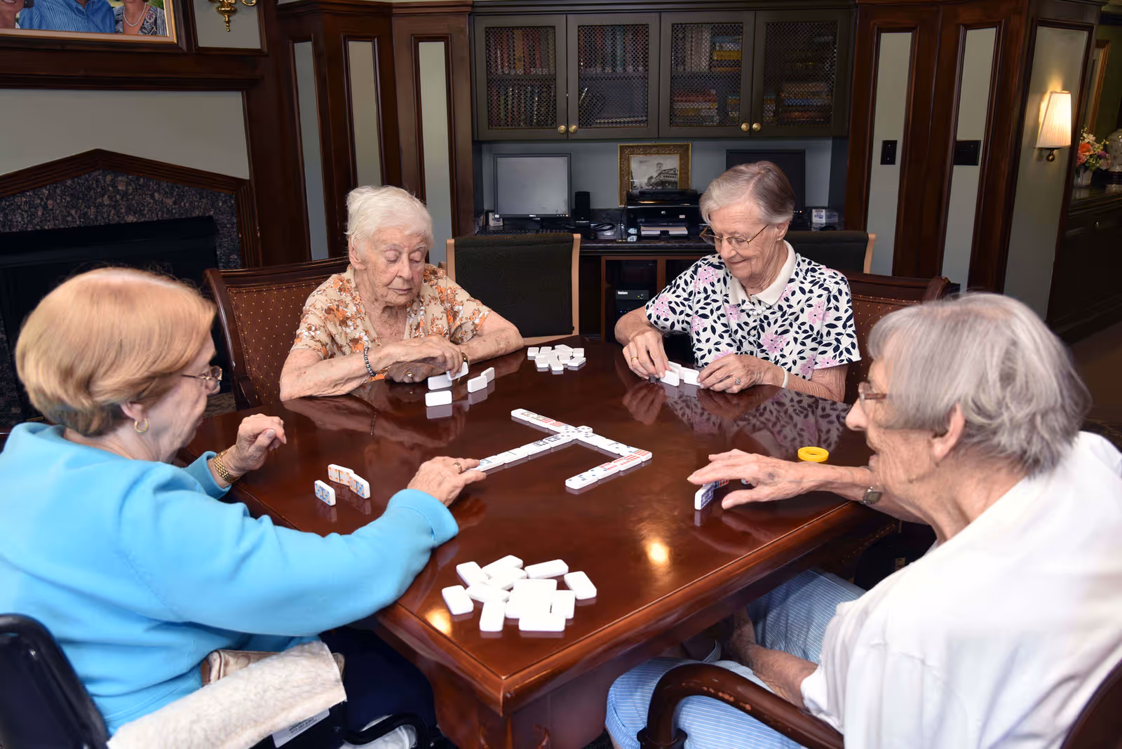Four elderly women sitting around a wooden table playing a game of dominoes in a cozy room with wooden furniture and a fireplace in the background.