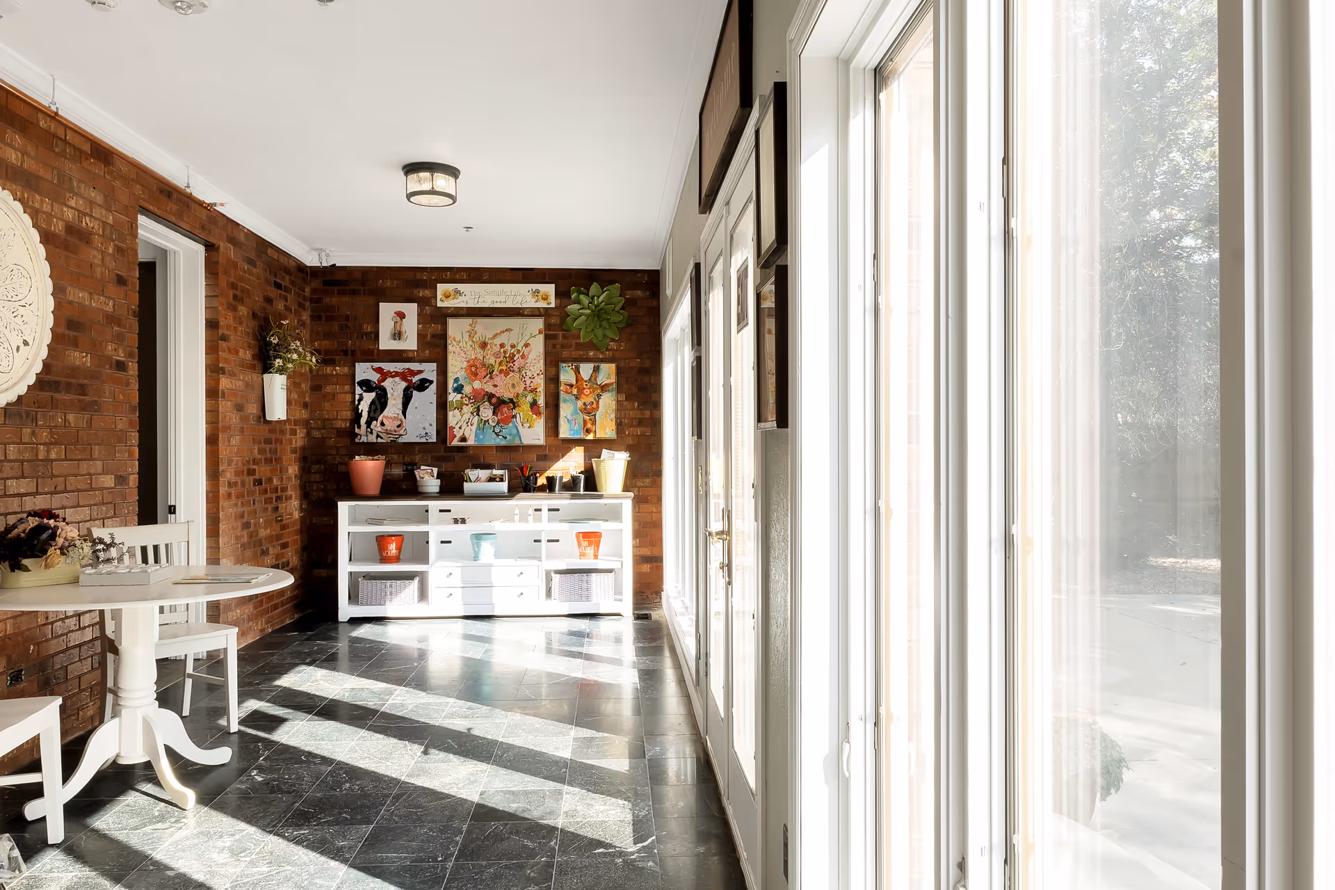 A bright interior hallway with large windows on the right letting in sunlight. On the left, there is a white round table with two chairs, and a white cabinet against a brick wall decorated with colorful animal and floral paintings. The floor is dark tile, and the space has a cozy, welcoming atmosphere.
