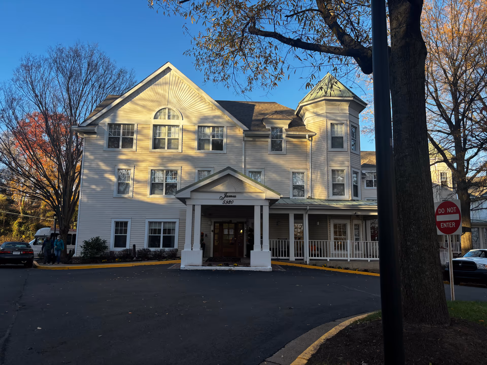 Exterior front view of a large, light-colored multi-story building with a covered entrance labeled 'James 5920'. The building has multiple windows, a turret on the right side, and is surrounded by trees with autumn foliage. There are cars parked on either side of the driveway and a 'Do Not Enter' sign near the right side of the image.