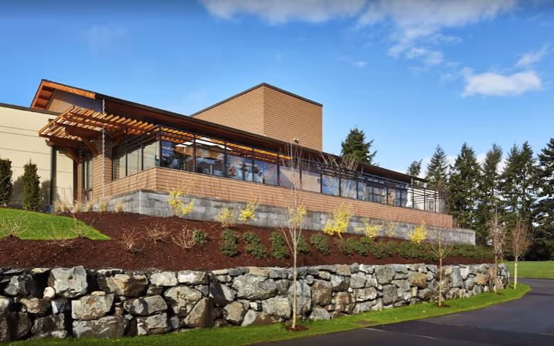 Modern single-story building with wood siding and large windows set above a stone retaining wall and landscaped grounds under a blue sky.
