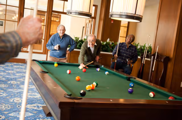 Three elderly men playing pool in a well-lit room with wooden paneling and large hanging lights above the pool table. One man is taking a shot while the other two watch and smile.