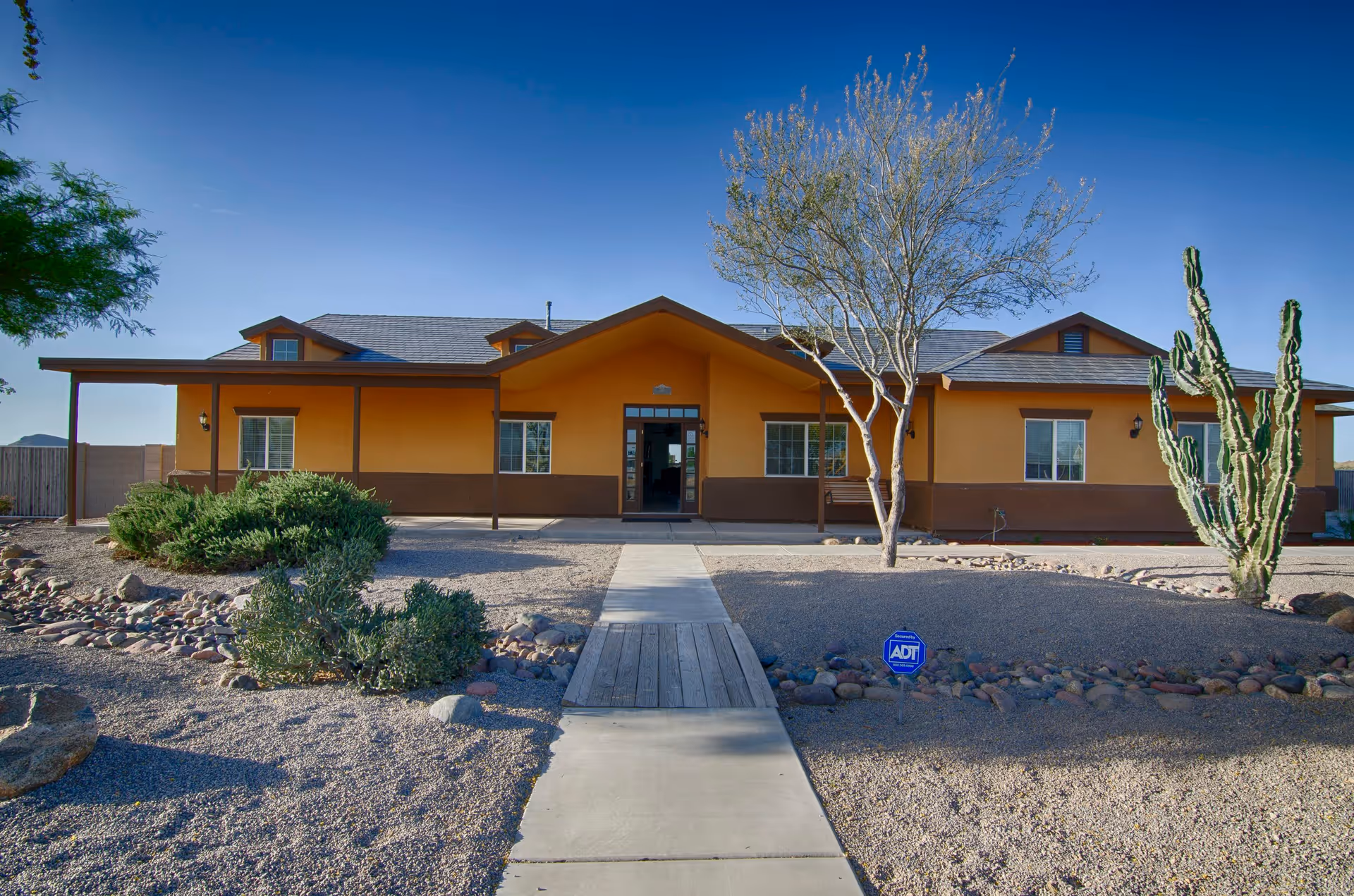 Single-story orange and brown building with a central entrance, a paved walkway, desert landscaping and cacti under a clear blue sky.