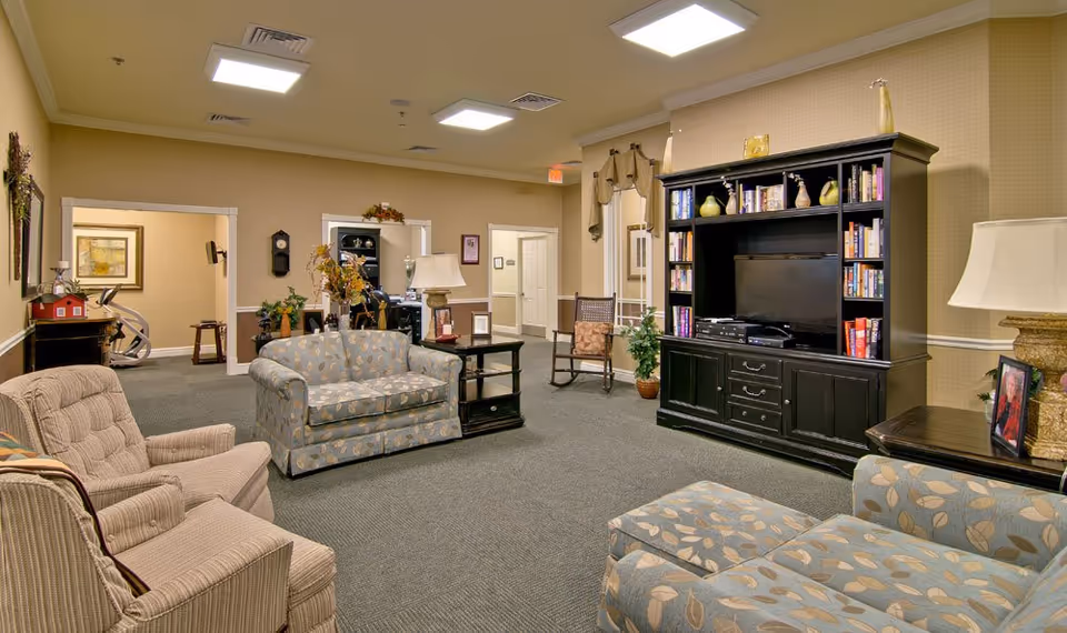 A cozy living room area in a senior living facility with patterned armchairs and a loveseat, a large black entertainment center with a TV and books, a rocking chair, side tables with lamps, and decorative plants. The room has beige walls, carpeted floors, and ceiling lights.