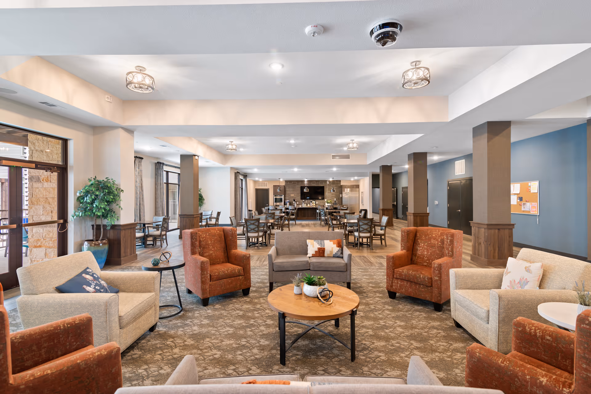 A spacious and well-lit common area in a senior living facility featuring comfortable seating with armchairs and sofas arranged around a wooden coffee table. In the background, there are multiple dining tables and chairs, a kitchen area with appliances, and large windows allowing natural light to enter. The room has a neutral color palette with blue and beige walls, carpeted flooring, and decorative plants.