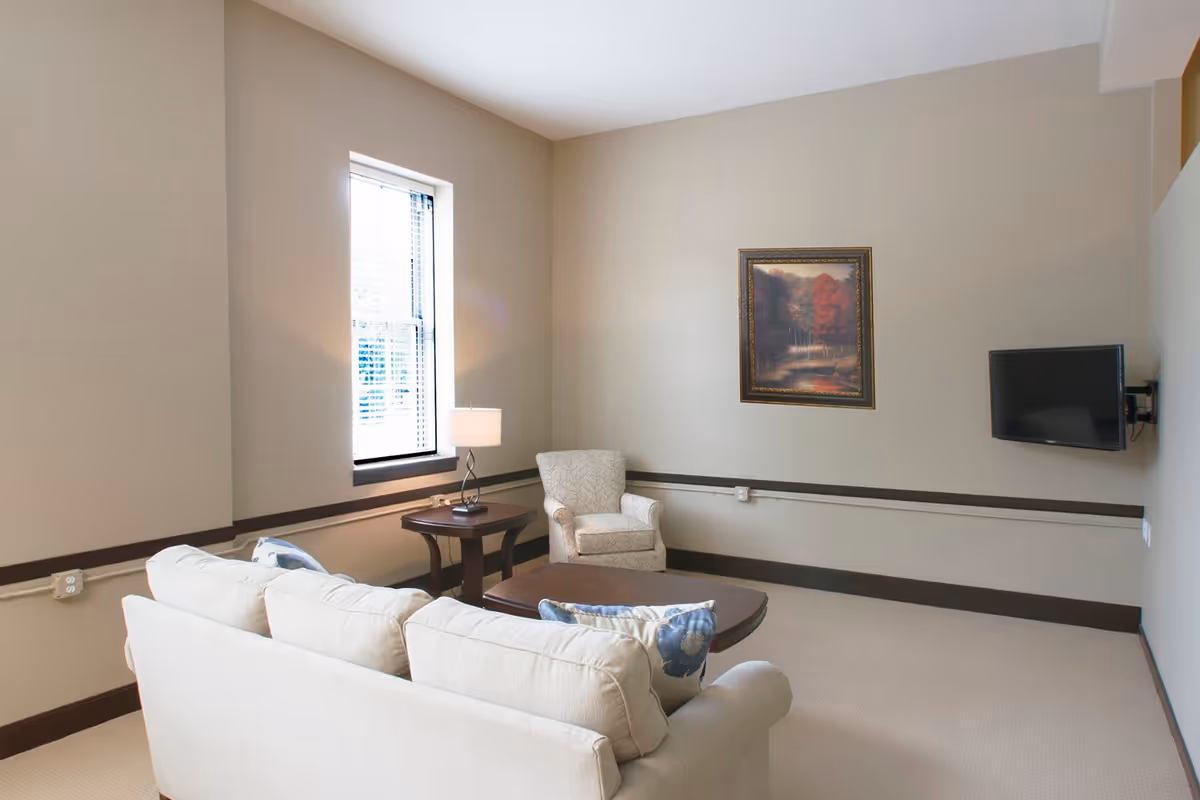 A cozy living room with a beige sofa adorned with blue and white floral pillows, a matching armchair, a wooden coffee table, and a side table with a lamp. There is a window with blinds letting in natural light, a framed painting on the wall, and a flat-screen TV mounted on the opposite wall.