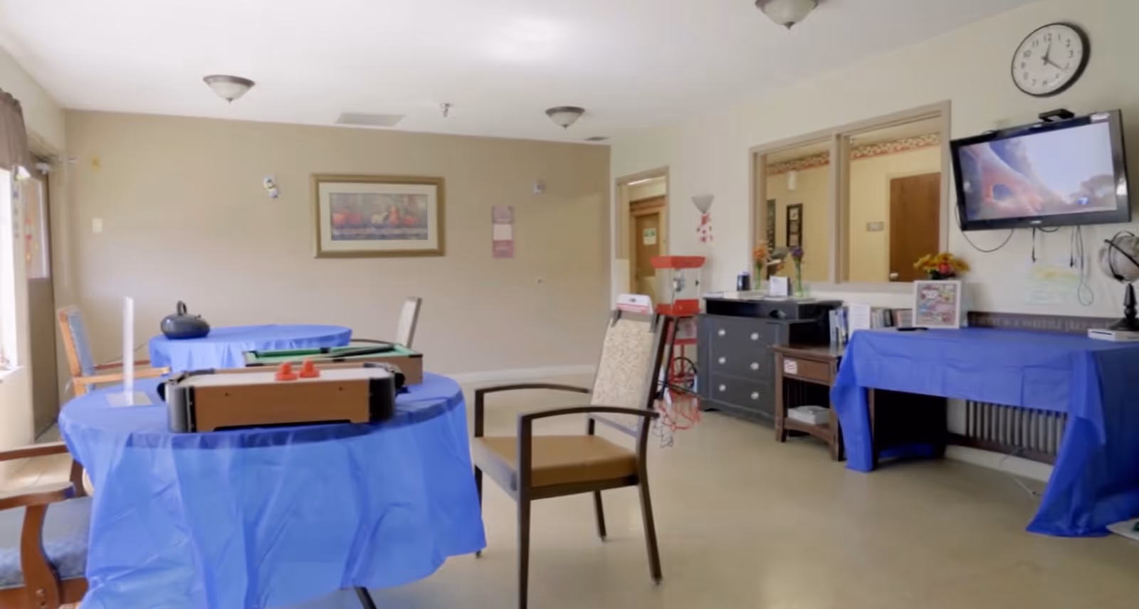 A common area in a senior living facility with tables covered in blue tablecloths, chairs, a wall-mounted TV, a clock, and various furniture including a dresser and a small table with a popcorn machine. The room has beige walls and ceiling lights.