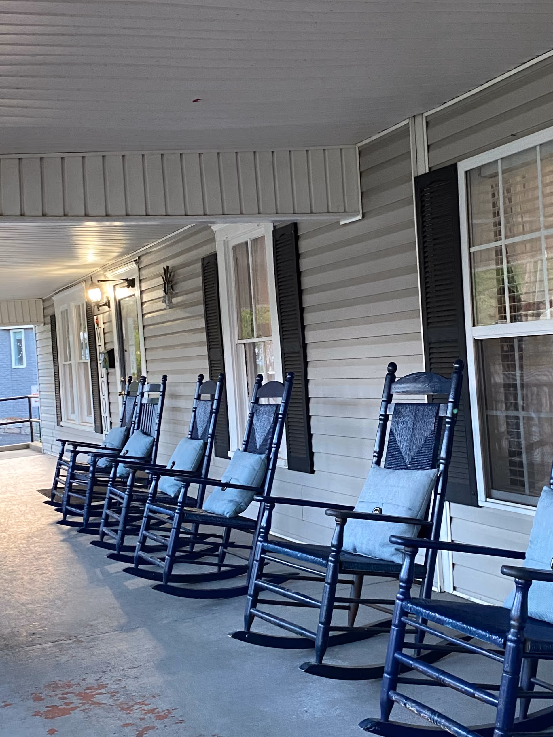 A covered porch area with a row of six dark blue wooden rocking chairs, each with a light blue cushion. The porch has beige siding with black shutters on the windows and a welcome sign near the door. The floor is concrete and the porch ceiling is white.