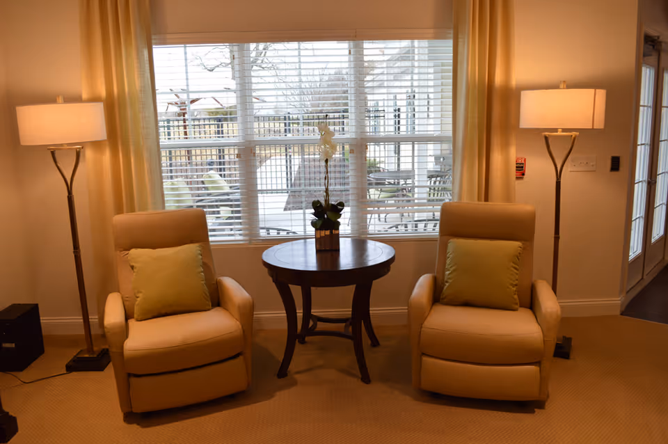 A cozy sitting area with two beige recliner chairs, each with a green cushion, positioned on either side of a round wooden table with a small potted plant. Behind the chairs is a large window with white blinds and light yellow curtains. Two floor lamps with beige shades stand on either side of the chairs, and a patio with outdoor furniture is visible through the window.