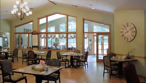 Dining room in a senior living facility with tables, chairs, large windows, and a clock.