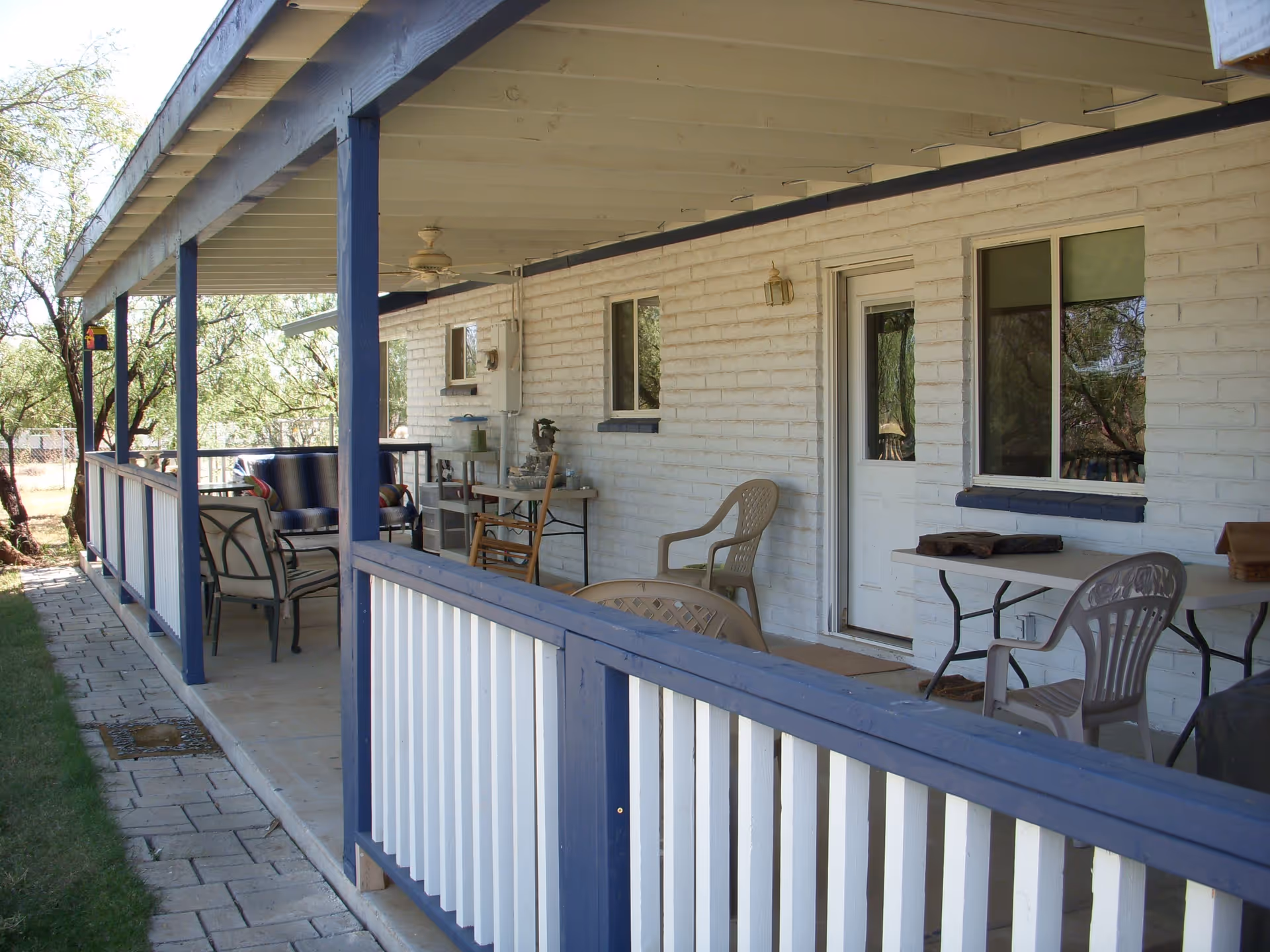 Covered outdoor patio area with white brick walls and blue trim, featuring various chairs, a table, and a ceiling fan, surrounded by trees and greenery.