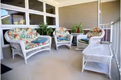 Covered outdoor patio with white wicker seating and colorful floral cushions, a glass-top wicker table, potted plants, and railing.