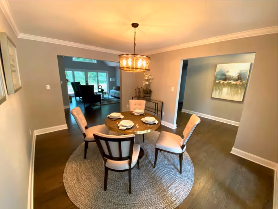 Dining room with a round table set for four under a chandelier, open to a living area.