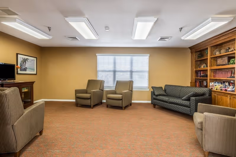 A cozy living room with beige walls and carpeted floor, featuring a window with blinds in the center. The room contains two beige armchairs, a patterned dark green sofa, and two additional beige armchairs arranged around the room. On the right side, there is a wooden built-in bookshelf filled with books and decorative items. On the left side, a small wooden cabinet holds a television and a framed picture hangs on the wall above it.