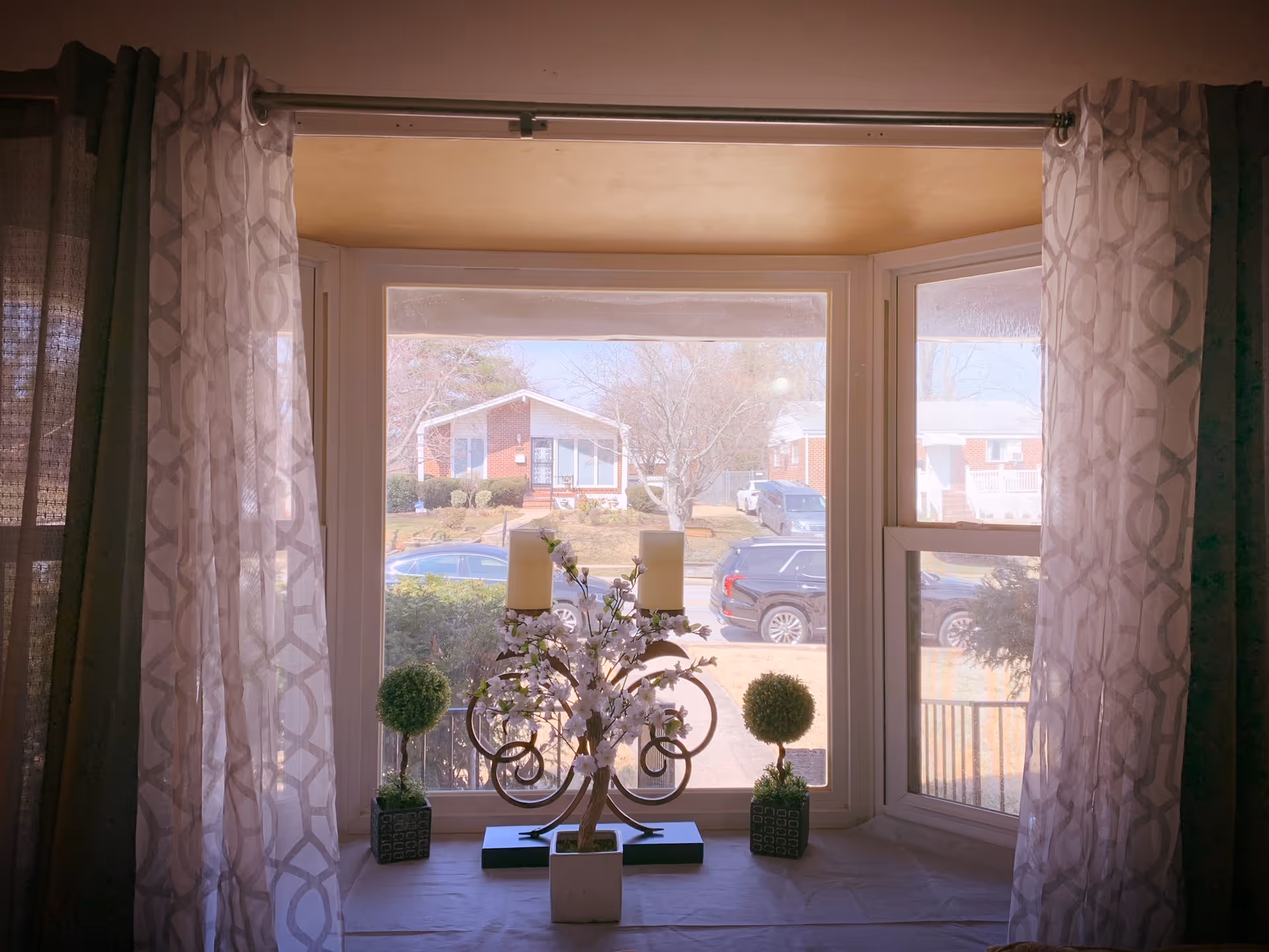 View from inside a living room through a bay window with patterned curtains and decorative candles and topiary on the windowsill, looking out to houses and parked cars.