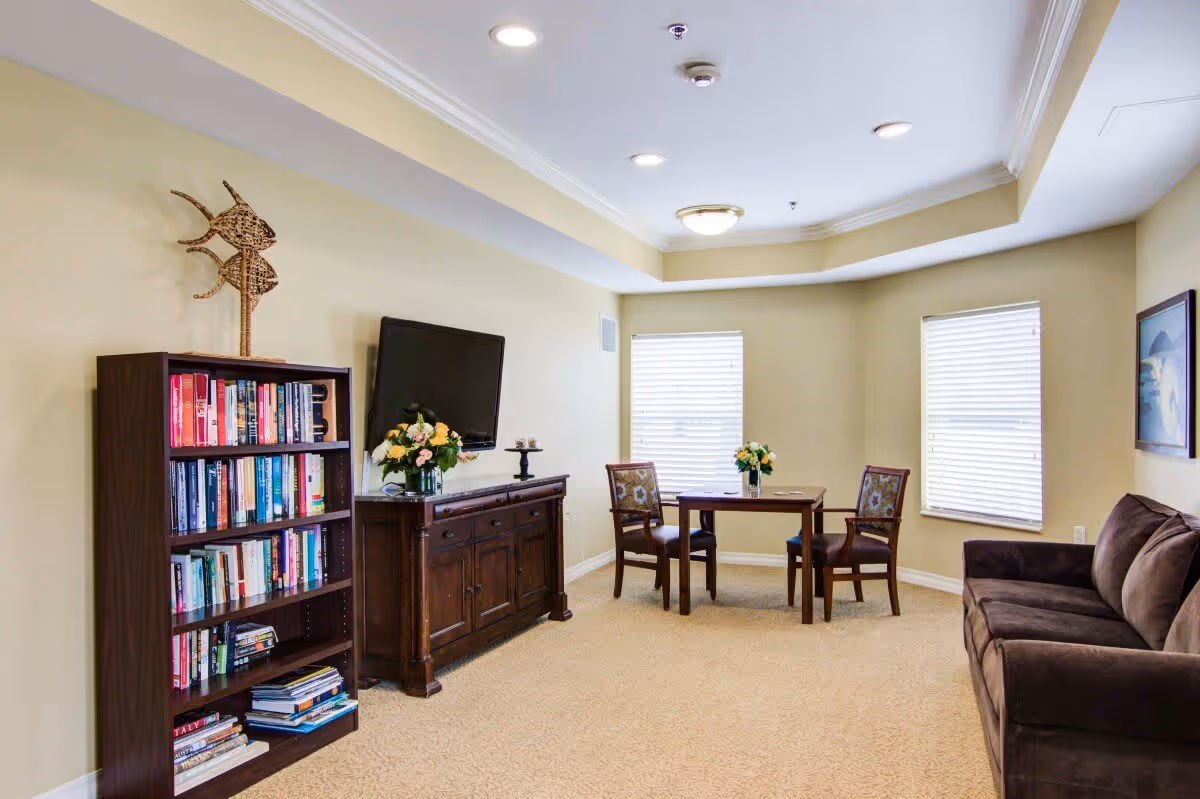 A cozy living room area with beige walls and carpet, featuring a dark brown sofa on the right, a wooden bookshelf filled with books on the left, a wooden cabinet with a flat-screen TV mounted above it, and a small wooden table with two chairs near two windows with blinds. There are flower arrangements on the cabinet and table, and a framed picture on the wall above the sofa.