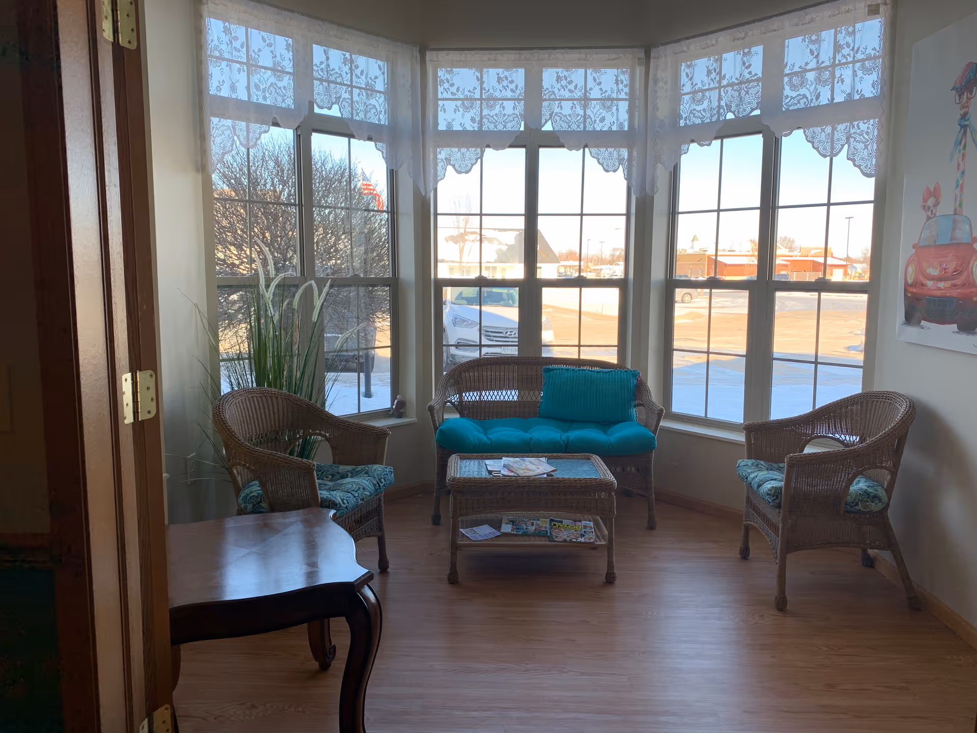 A cozy sitting area with three wicker chairs and a wicker loveseat, all with blue-green cushions, arranged around a wicker coffee table with magazines on it. The space is well-lit by large windows with lace curtains, showing a view of a parking lot and some trees outside. There is a tall plant in the corner and a colorful framed artwork on the wall to the right.