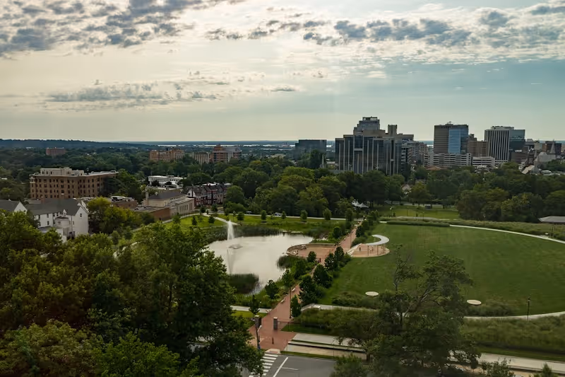 Aerial view of a city park with a pond featuring a fountain, surrounded by trees and walking paths. In the background, there is a city skyline with multiple tall buildings under a partly cloudy sky.