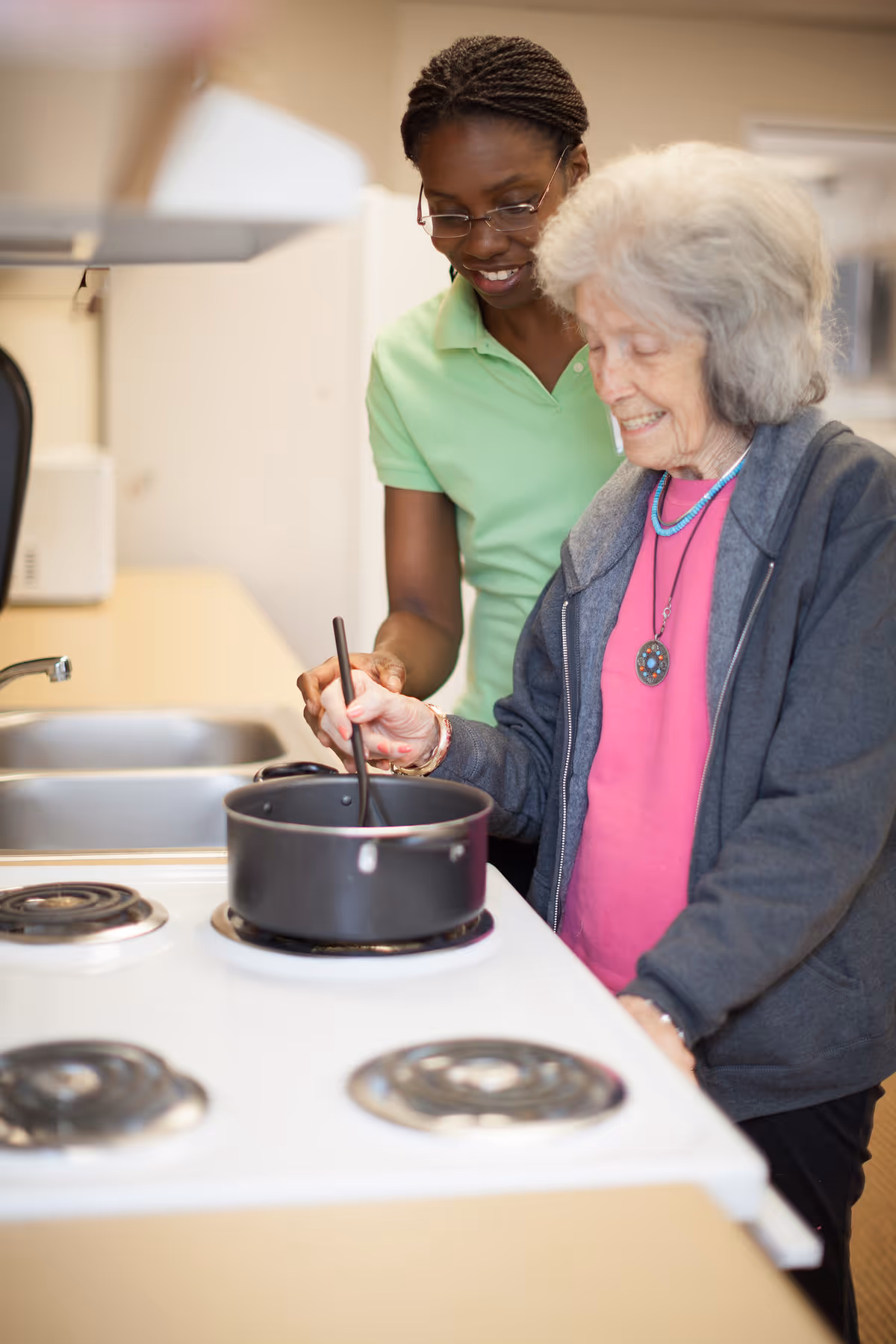 An elderly woman stirring a pot on a stove with the assistance of a smiling caregiver in a kitchen setting.