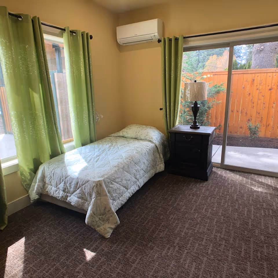 A small bedroom with a single bed covered in a light-colored quilt, green curtains on two windows, a dark wooden nightstand with a lamp, and a sliding glass door leading to an outdoor area with a wooden fence and trees.