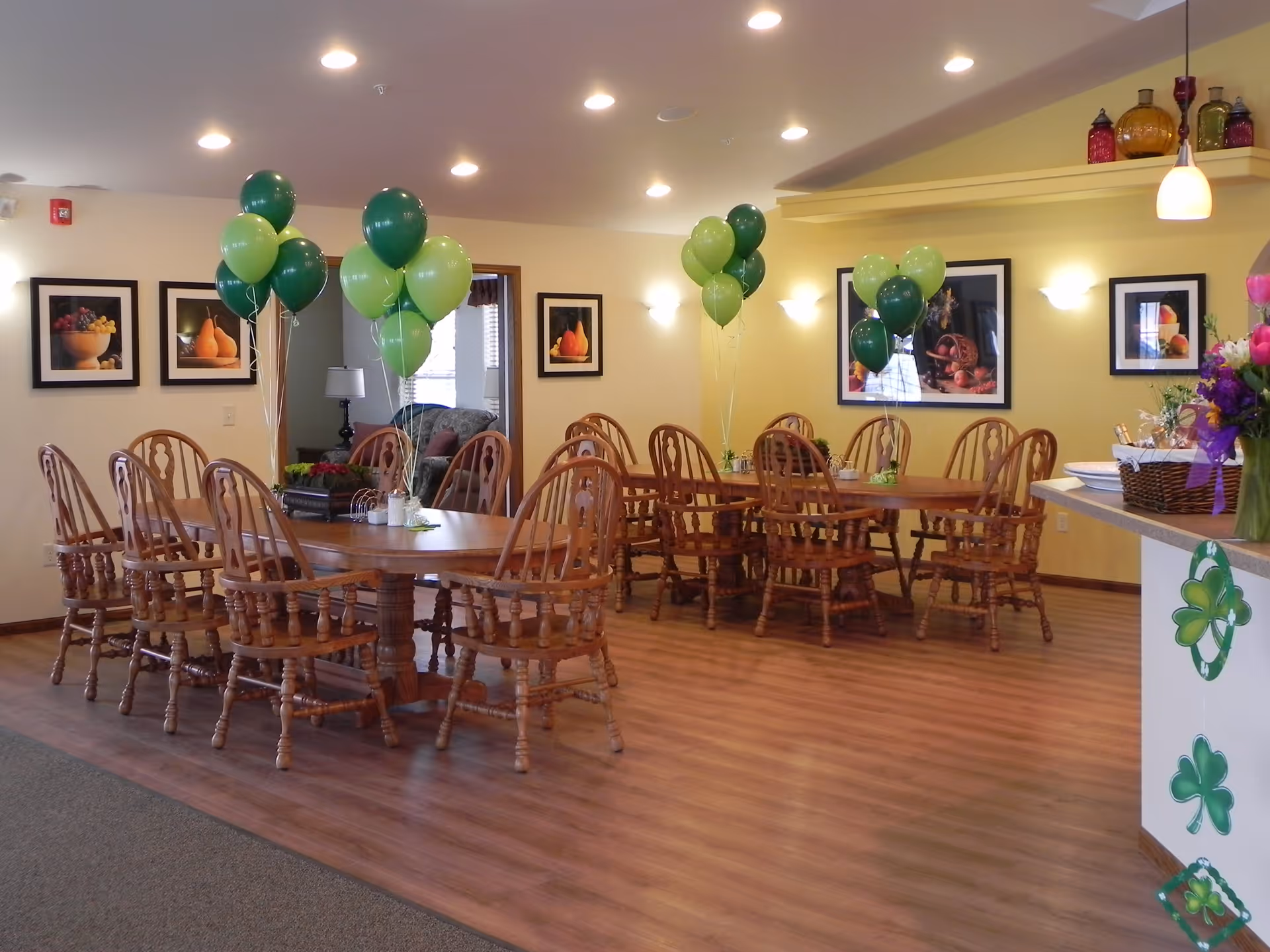 A dining room with wooden tables and chairs arranged neatly. Green and light green balloons are tied to the tables. The walls are decorated with framed pictures of fruits, and there is a counter on the right side with floral decorations and shamrock cutouts hanging from it. The room has wooden flooring and recessed ceiling lights.