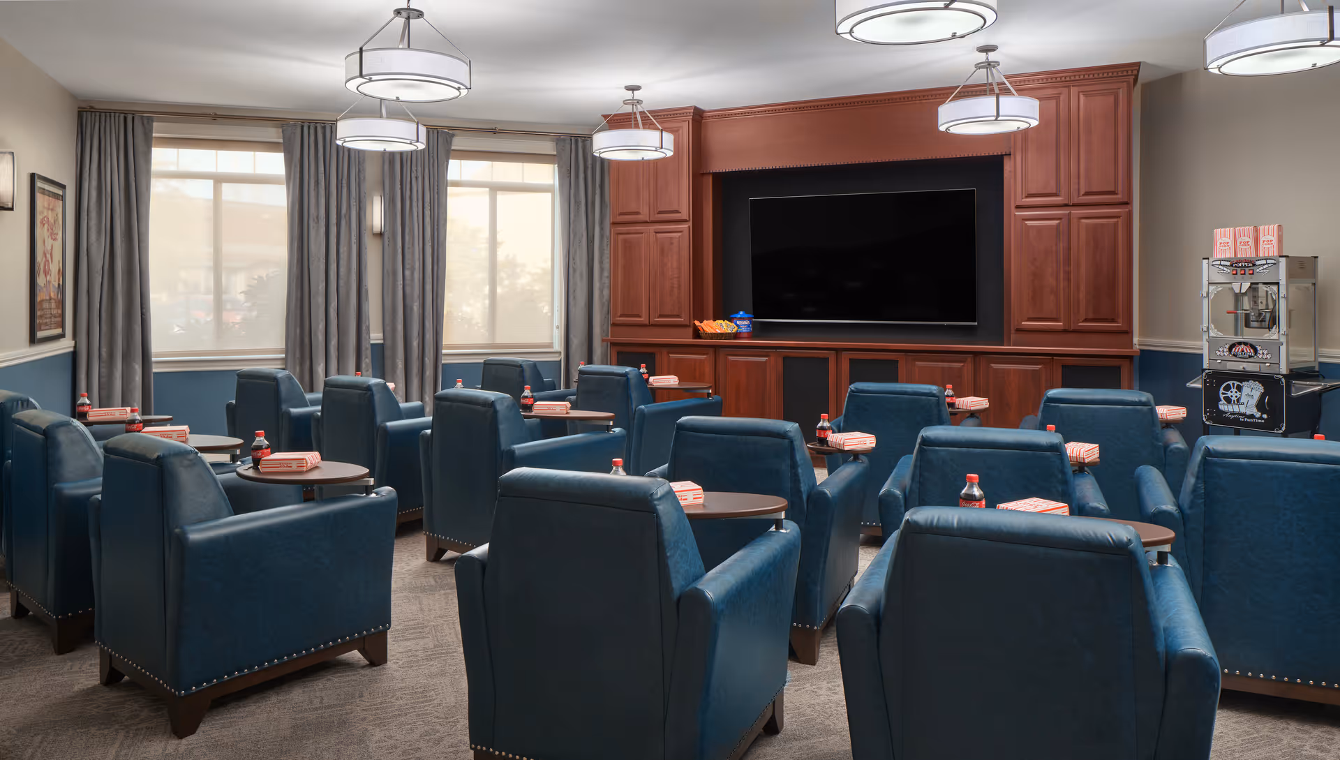 A senior living facility media room with multiple blue leather armchairs arranged in rows facing a large flat-screen TV mounted on a wooden entertainment center. Each chair has a small round table attached, holding a bottle of Coca-Cola and a striped popcorn box. There is a popcorn machine in the corner of the room, and large windows with gray curtains let in natural light.