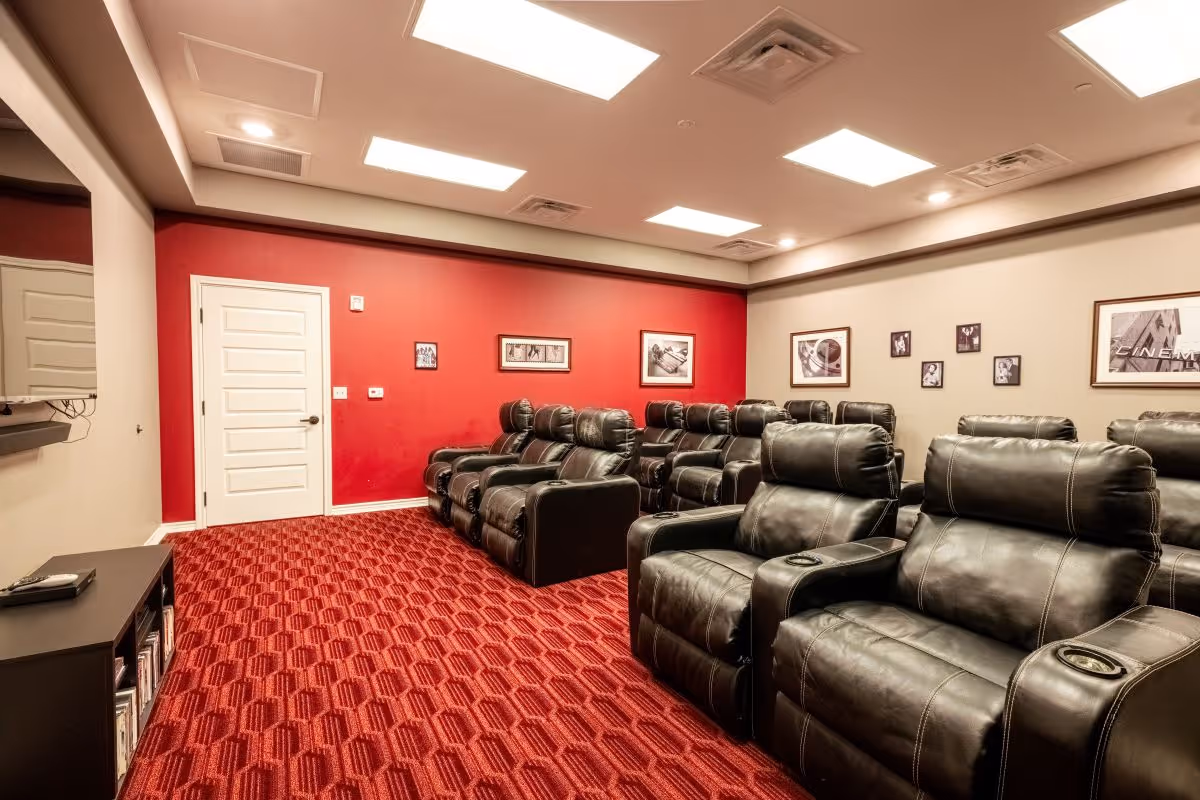 A cozy home theater room with multiple rows of black leather recliner chairs arranged on a red patterned carpet. The walls are painted beige and red, adorned with framed black and white pictures. The ceiling has recessed lighting panels.