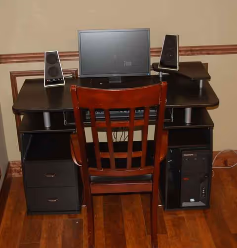 Dark wooden desk with monitor, two speakers, and a wooden chair in an interior room.