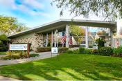 Entrance of a one-story rehabilitation building with a stone facade, American flags, manicured lawn, and a sign for Palo Alto Sub-acute and Rehabilitation Center.