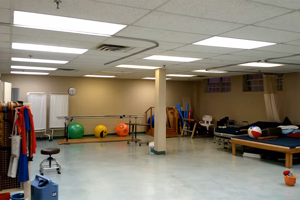 A spacious therapy or rehabilitation room with exercise balls, parallel bars, therapy beds, and various physical therapy equipment. The room has a tiled ceiling with fluorescent lighting and beige walls.