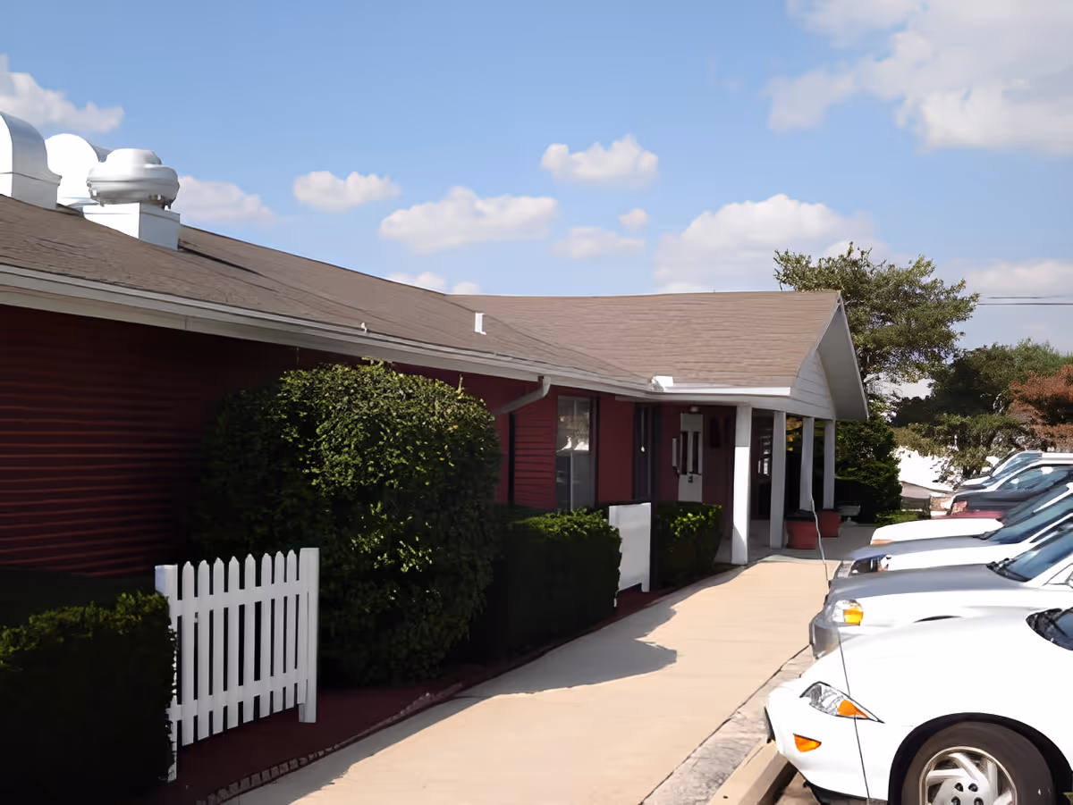 Exterior view of a single-story building with a brown roof and red siding, a white picket fence, trimmed bushes, a sidewalk, and several parked cars under a partly cloudy sky.