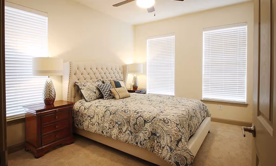 A well-lit bedroom with a large bed featuring a tufted headboard and paisley-patterned bedding. There are two wooden nightstands on either side of the bed, each with a decorative lamp. The room has three windows with white blinds and beige walls.