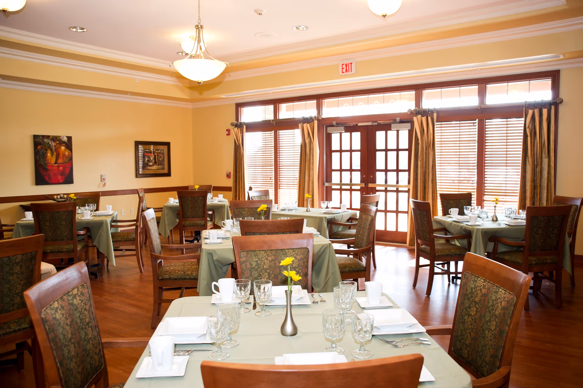 A well-lit dining room with multiple tables covered in light green tablecloths, each set with glassware, white plates, and silverware. Wooden chairs with patterned cushions surround the tables. The room has large windows with wooden blinds and curtains, and two framed paintings on the wall. Ceiling lights provide warm lighting.