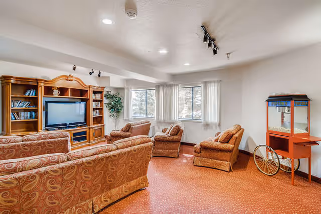 A cozy living room with patterned upholstered sofas and armchairs arranged around a wooden entertainment center with a flat-screen TV. There are two windows with white curtains letting in natural light, a green potted plant in the corner, and a vintage-style popcorn machine on wheels against the wall.