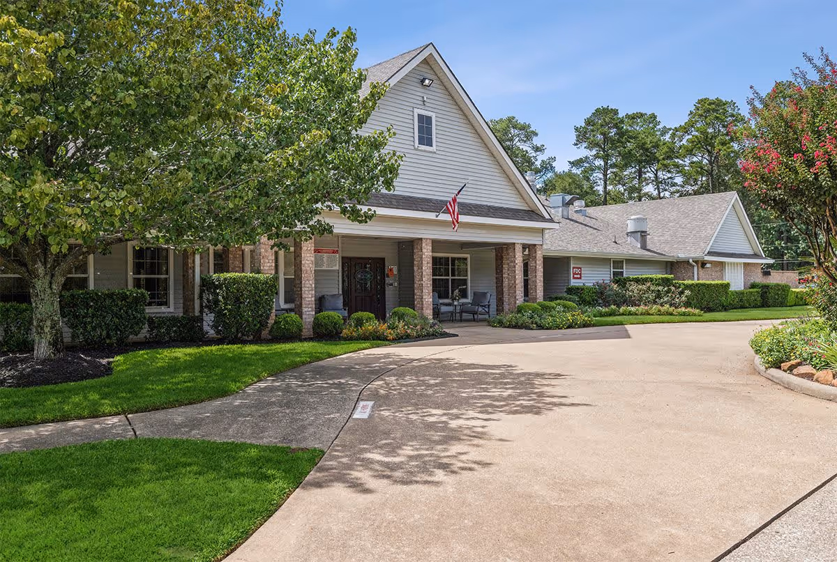 Exterior view of Strake Court facility showing a driveway leading to the entrance with a covered porch supported by brick columns. The building has light gray siding with a peaked roof, surrounded by well-maintained green lawns, bushes, and trees under a clear blue sky.