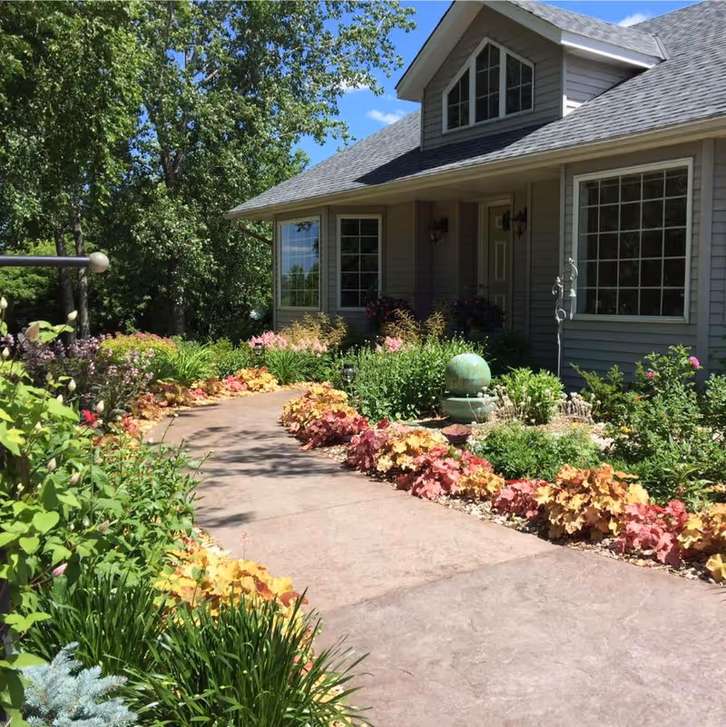 A paved walkway through colorful landscaped gardens leading to the front entrance of a house with large windows.