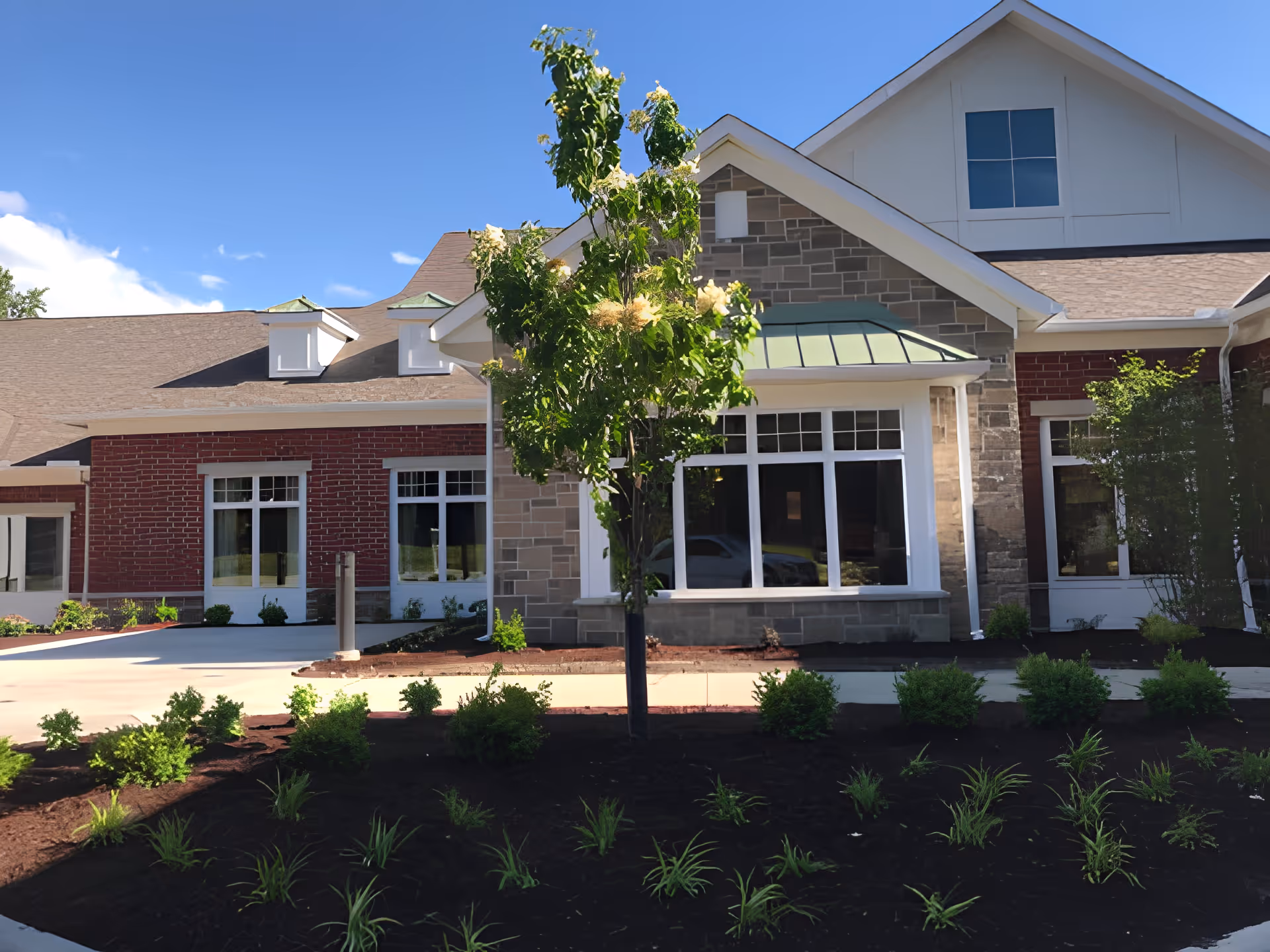 Exterior view of a senior living facility building with a mix of brick and stone facade, large windows, a small tree, and landscaped garden beds in front under a clear blue sky.